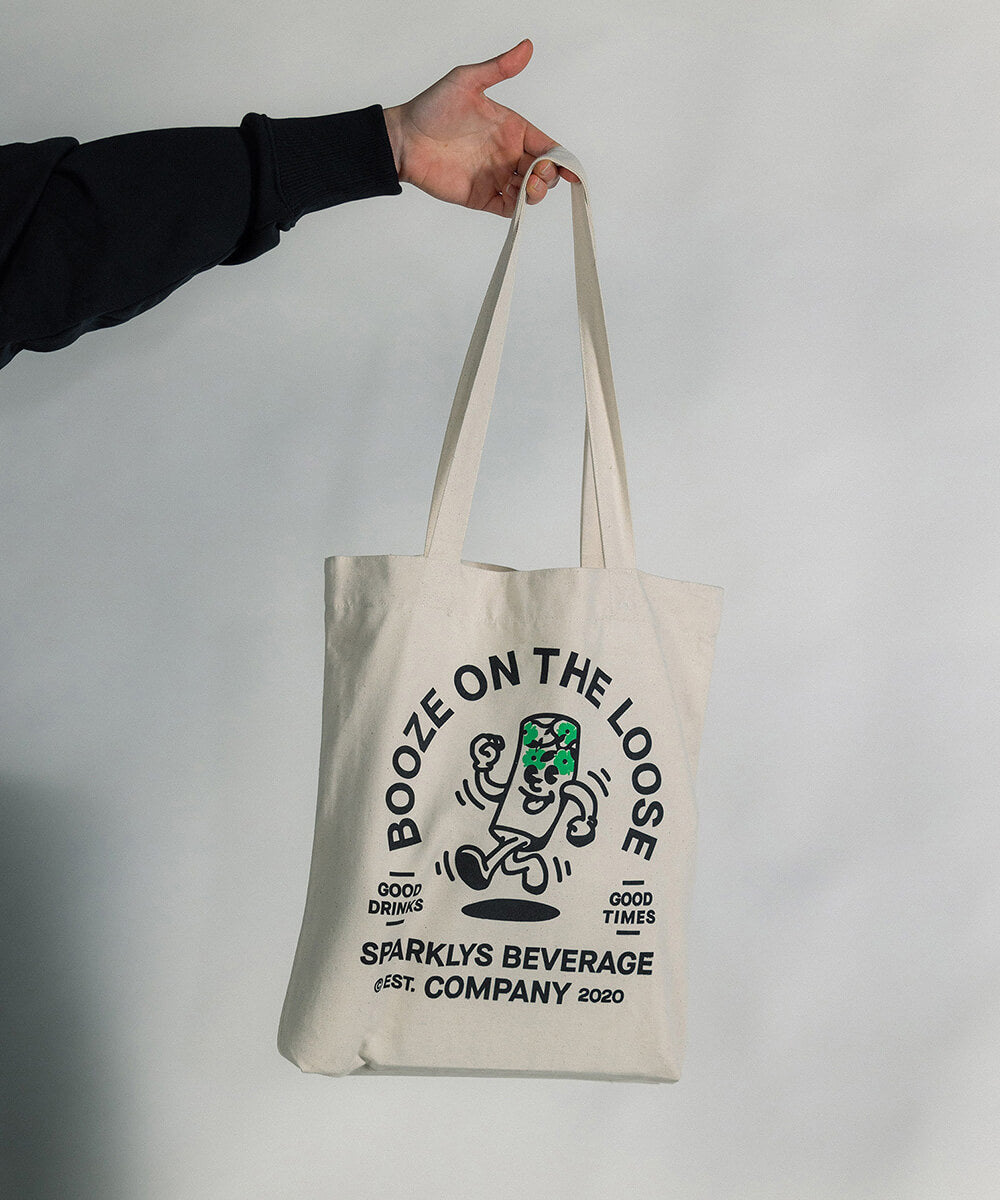 Eine Hand hält den Sparklys Tote Bag in der Luft, zeigt die Rückseite mit der Vintage-Comic-Stil Grafik von Sparklys Hard Seltzer Holunder Dose und dem Slogan 'Booze on the loose'.