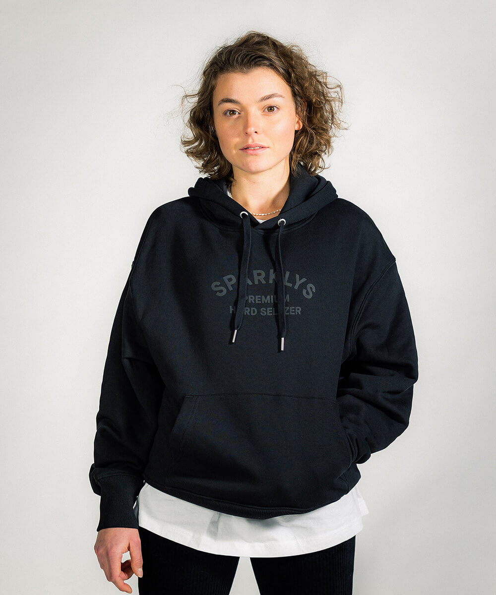 Weibliches Model präsentiert die Vorderseite des Nutrition Facts Hoodies von Sparklys, mit dem subtilen, in Schwarz gedruckten Vintage-Logo von Sparklys Hard Seltzer auf der Brust.